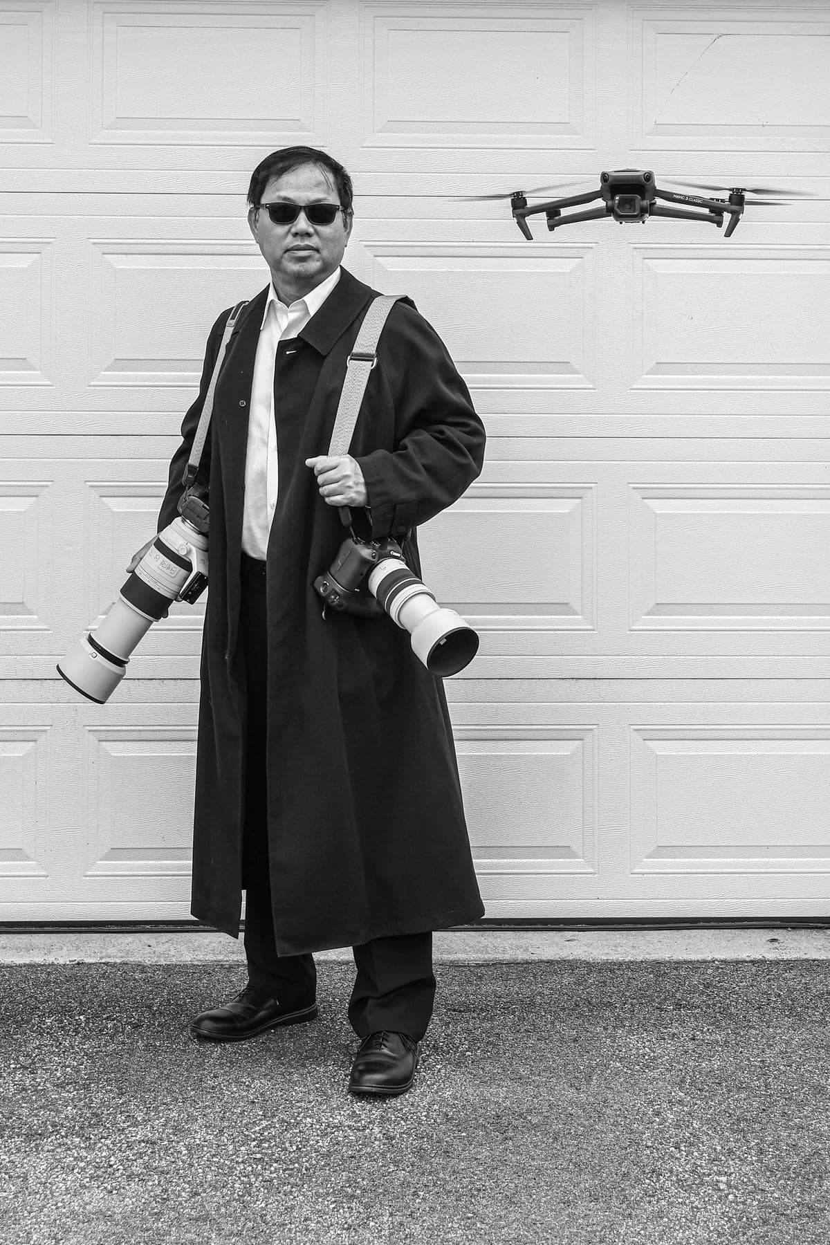 Carlton Soohoo in a long dark coat, a Canon telephoto over each shoulder and a DJI drone hovering beside him, in black and white