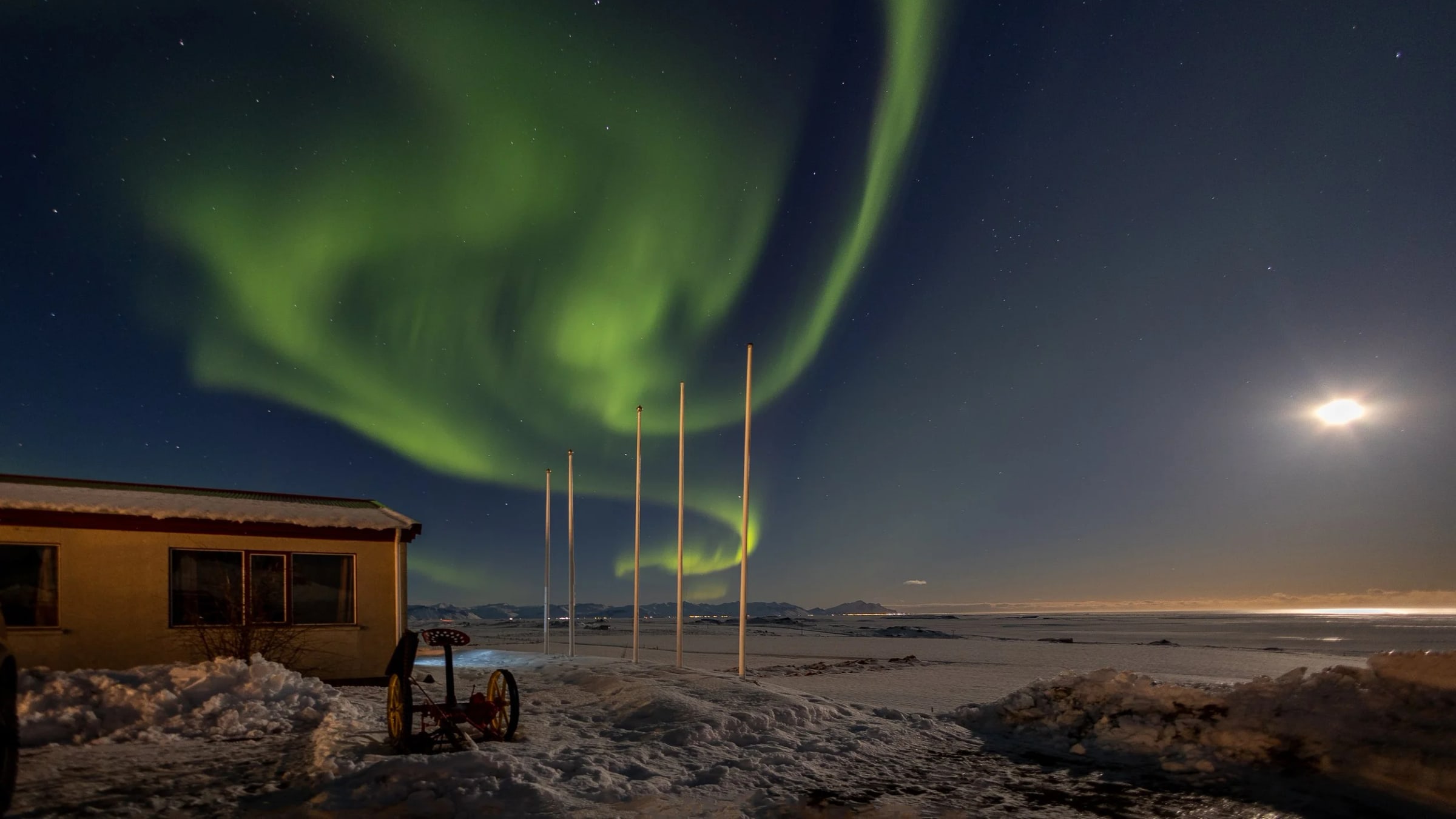 Green aurora borealis curling across the night sky above a lit cabin with the moon low on the horizon, by Carlton Soohoo